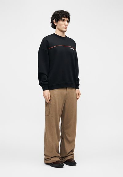 Man met krullend haar die een zwarte trui met een rode lijn en tekst draagt, losse bruine broek en donkere schoenen, staande tegen een witte achtergrond.