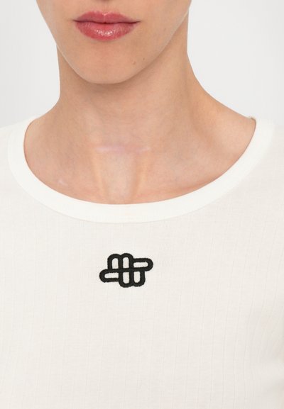 White ribbed fabric top featuring a round neckline and a black embroidered detail resembling a knot at the chest area.
