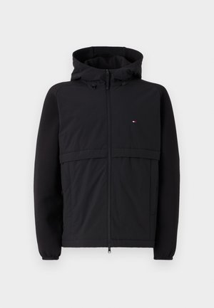 Schwarze Jacke mit Reißverschluss vorne, Kapuze und kontrastierenden Materialien sowie einer strukturierten Frontplatte. Auffälliges kleines Logo auf der Brust.