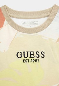 T-shirt in cotone biologico con base beige, caratterizzato da motivi floreali astratti in tonalità pastello. Ricamo nero "GUESS EST. 1981" sulla parte anteriore.