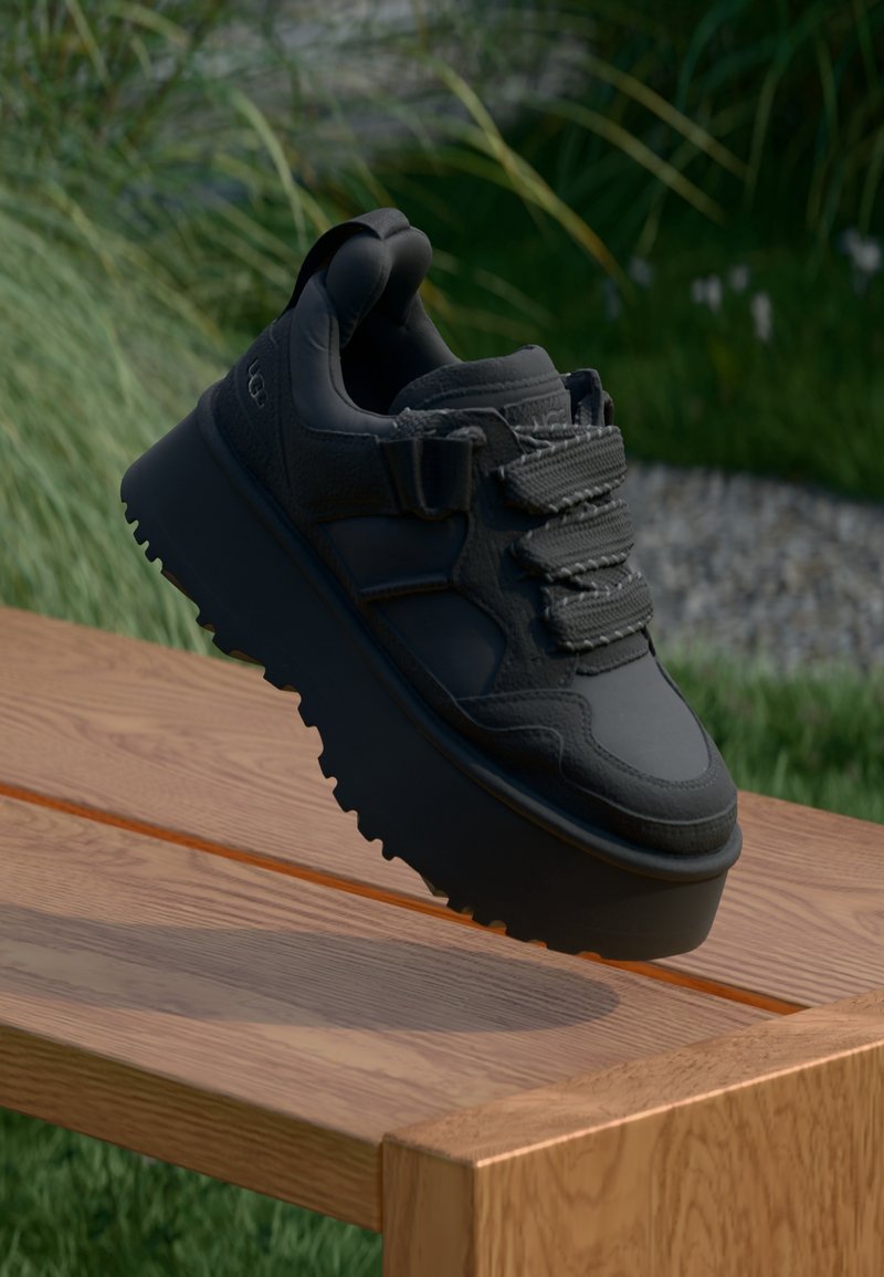 Svart sneaker med texturerad ovansida, tjock sula, lager-på-lager-design och dekorativa snören, visad mot en träyta.