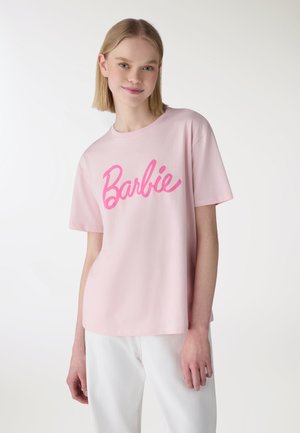 Giovane donna che indossa una maglietta rosa chiaro con la scritta "Barbie" in corsivo rosa brillante, abbinata a pantaloni bianchi, in piedi davanti a uno sfondo semplice.