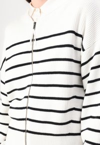 Biały sweter dzianinowy w poziome czarne paski, z zamkiem z przodu, wysokim kołnierzem i teksturowanym materiałem. Akcentowany prążkowanymi detalami.