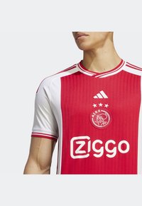 Mężczyzna w czerwono-białej koszulce piłkarskiej Ajax Amsterdam z logo Adidas i sponsorem Ziggo na piersi.