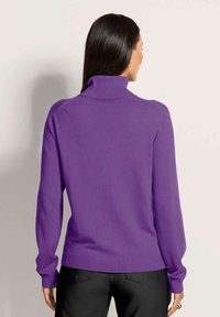 Frau mit langen dunklen Haaren, die einen lila Rollkragenpullover und schwarze Hosen trägt und mit dem Rücken zugewandt steht.