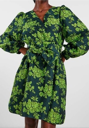 Woman wearing dark green dress with bright green floral pattern, puffed long sleeves, V-neck, and a large bow tied at the waist.