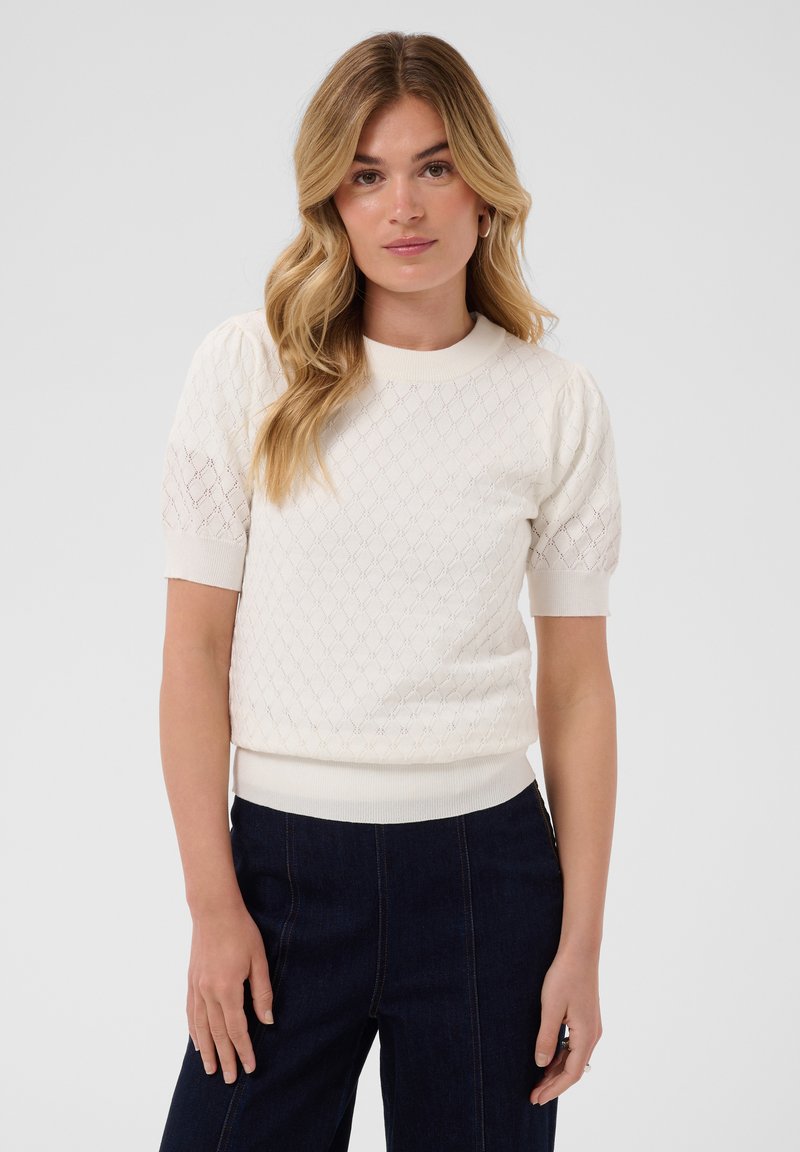 Femme blonde aux cheveux ondulés portant un pull texturé blanc à manches courtes et un pantalon sombre taille haute sur un fond uni.