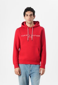 Rød bomullshoodie med frontlomme, snor i hetten, og trykket logo "TOMMY HILFIGER" med "EST. 1985" under. Ledig passform, ribbet mansjetter.