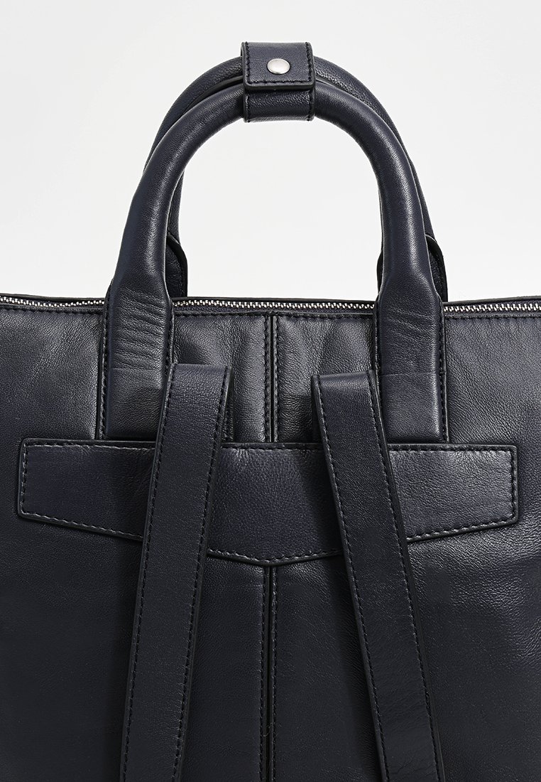 Czarna skórzana torba na ramię z okrągłymi uchwytami, zamkiem błyskawicznym i płaskimi paskami z detalami szewnymi na zewnętrznej części. Gładka tekstura.