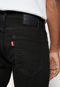 Gros plan d'une personne portant un jean noir Levi's avec une poche arrière visible, l'onglet rouge Levi's, un patch de marque en cuir noir et une partie d'une chemise blanche.