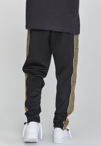 Pantalones deportivos negros con franjas laterales doradas y una textura suave, combinados con zapatillas blancas que tienen detalles texturizados y cordones.