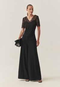 Črna čipkasta majica s skledastim izrezom skupaj s širokimi črnimi hlačami. Model drži črno torbico. Gladek material, eleganten dizajn.