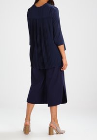 Tunic plissettato blu navy con maniche a tre quarti, abbinato a culotte blu navy con spacchi laterali. Indossato con sandali con tacco a blocco beige.