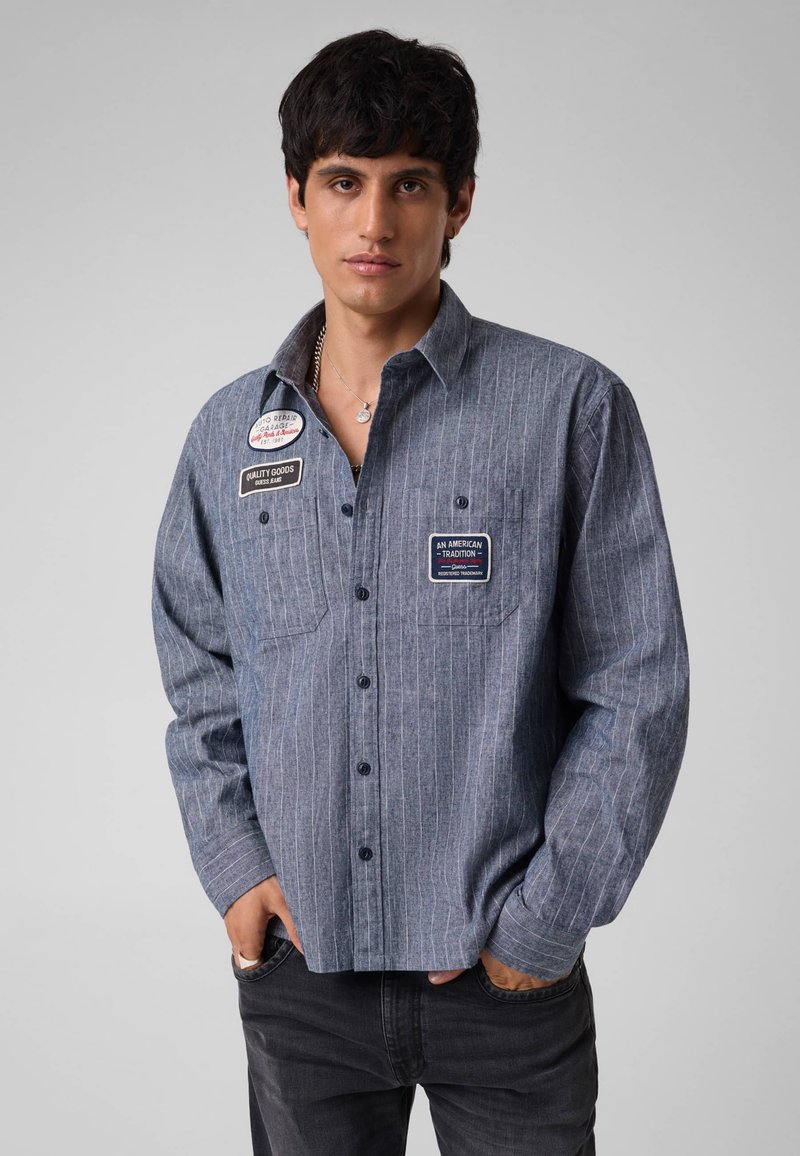 Jeune homme aux cheveux foncés portant une chemise bleue à fines rayures avec des patchs et un jean noir, debout les mains dans les poches.