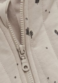 Lys beige stof med en glat tekstur, der har små mørke træ- og poteaftryksmønstre. Inkluderer en beige lynlås med en rund trækknap.