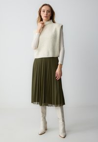 Top senza maniche in maglia color avorio, abbinato a una gonna plissettata verde oliva e stivali bianchi al ginocchio, che mette in risalto una texture liscia e una silhouette moderna.