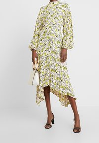 Femme portant une robe longue à manches longues, dégradée, à fleurs jaunes, tenant un sac à main blanc avec des poignées en bois, et des sandales à talons noires.