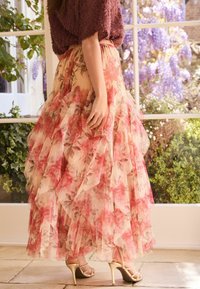 Maxi-Rock mit Blumenmuster aus weichem, durchsichtigem Stoff mit gerüschten Lagen in Rosa- und Pfirsichtönen, kombiniert mit goldenen Riemchenabsätzen.