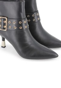 Botas de tobillo de cuero negro con punta afilada, herrajes en acento dorado, y una correa ancha que presenta recortes circulares y una hebilla.