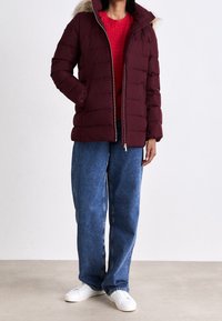 Bordowa kurtka puchowa z futrzanym kapturem, z suwakiem i pikowanym wzorem. Zestawiona z niebieskimi, luźnymi dżinsami i białymi trampkami.