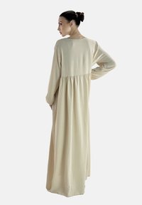 Lange, beige jurk van zachte stof met een ontspannen pasvorm, voorzien van lange mouwen en een gedetailleerde taille met plooien. Soepele textuur en eenvoudig ontwerp.