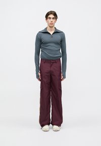 Prilegajoča siva majica z dolgimi rokavi in zadrgo na ovratniku, v kombinaciji z širokimi smetanastimi cargo hlačami, ki imajo stranske žepe in rumene črte. Masivni beli čevlji.