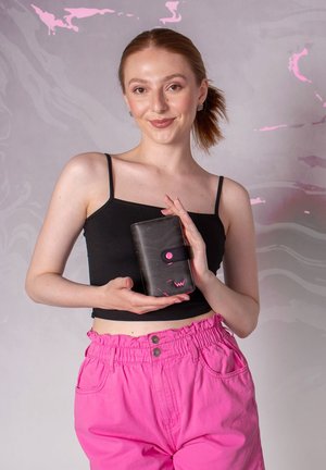 Woman with light skin and red hair in a ponytail holding a black wallet with pink accents, wearing a black top and bright pink pants.