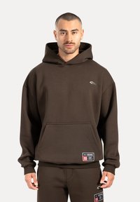 Brauner Hoodie aus weichem Material mit einer großen Vordertasche, gerippten Bündchen und einem kleinen Logo auf der Brust. Hat einen lässigen Schnitt und passende Hosen.