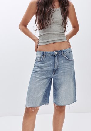 Nainen, jolla on vaaleansiniset denimin Bermudan shortsit, joissa on pieniä valkoisia maaliroiskeita, ja harmaa hihaton ribbineuletoppi, kädet lanteilla.