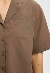 Kurze Ärmel, braune Bluse aus glattem Stoff; mit Kragen und einer aufgesetzten Tasche auf der linken Seite. Knopfleiste vorne.