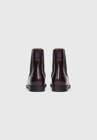 Une paire de bottes Chelsea en cuir brun foncé, poli, vues de l'arrière, avec des panneaux latéraux élastiques noirs et des talons bas noirs sur un fond blanc.