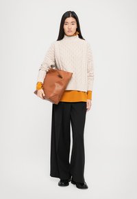 Chunky beige cable knit sweater, rust turtleneck underneath, loose black trousers, and a large brown leather bag. Model standing against a white background.