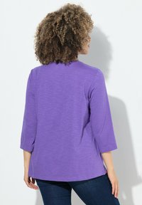 Lila, topp med 3/4 ärm i mjukt tyg. Har en avslappnad passform och rundad nederkant, med en jämn yta och minimal mönstring.