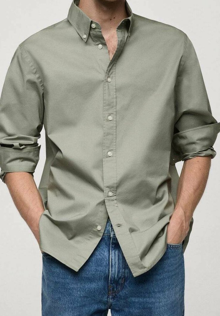 Homme portant une chemise boutonnée vert clair avec manches retroussées et un jean bleu, mains dans les poches, sur fond uni.