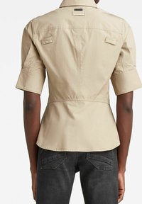 Beige skjorta med kort ärm, figurnära midja och ryggsömn. Har två bröstfickor och rundad nederkant. Tillverkad av ett lätt tyg.