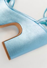 Vaaleansininen korkokenkä, jossa on metallinen pinta, avonainen muotoilu ja paksu lohkopohja. Sisältää ristikkäin olevat hihnat ja pyöreän varvasosan.