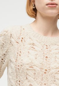 Γυναίκα που φοράει πουλόβερ ανοιχτής πλέξης σε κρεμ απόχρωση με σχέδια καλωδίων και στρογγυλή λαιμόκοψη, δείχνοντας μέρος του προσώπου και του λαιμού της.