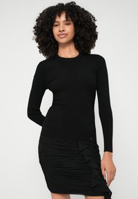 Fekete bordázott, hosszú ujjú felső, testhez simuló sziluettel, párosítva egy fekete redőzött szoknyával, amelynek az oldalán fodrok díszelegnek.