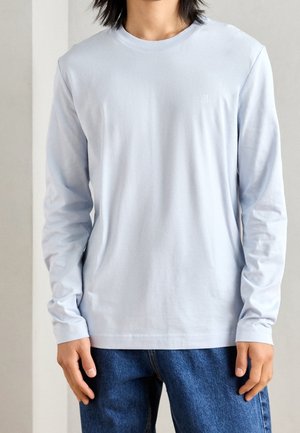 Person iført en ensfarvet lyseblå langærmet skjorte og blå jeans, stående med hænderne afslappet ved siderne mod en neutral baggrund.