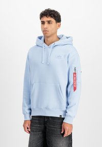 Világoskék kapucnis pulóver, puha anyagból, elülső zsebből, húzózsinóros kapucnival és piros kiegészítő címkével a ujjon.