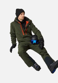 Zielony kombinezon narciarski z pomarańczowymi akcentami, czarna czapka beanie, rękawice i buty narciarskie. Trzyma czarny kask z niebieskim wizjerem. Teksturowany materiał, casualowy design.