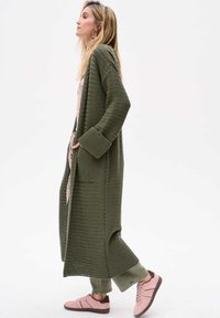 Olijfgroene gebreide cardigan met lange mouwen en twee zakken, gedragen over een bloemen-top en lichtgroene broek, gecombineerd met roze sneakers.