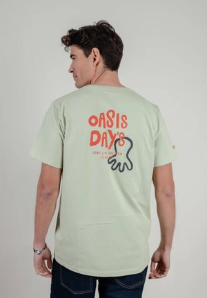 Hombre con camiseta verde claro con texto rojo "OASIS DAY'S" y gráfico abstracto de mano negra en la espalda, de espaldas a la cámara.