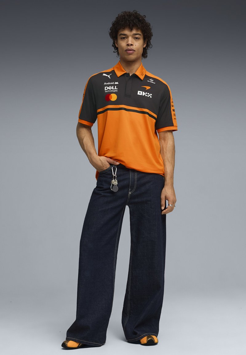 Joven de pie con polo del equipo de carreras en naranja y negro con logotipos de patrocinadores, jeans oscuros de pierna ancha y zapatillas deportivas naranjas, con llaves sujetas al jeans.