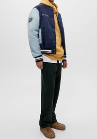 Person som bär en marinblå och ljusblå varsityjacka med texten "Yale University", en senapsgul hoodie, mörka byxor och bruna avslappnade skor, står upp.