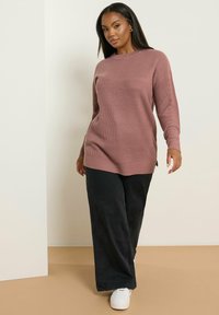 Pull en tricot rose à côtes longues avec fentes sur les côtés, associé à un pantalon large noir et des baskets blanches. Texture douce et coupe décontractée.