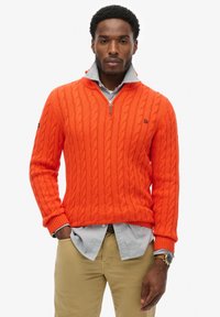 Pomarańczowy sweter z dzianiny warkoczyków z pół-zamkiem, noszony na jasnoniebieskiej koszuli z guzikami. Posiada ściągacze na rękawach i u dołu. Prosty detal z logo.