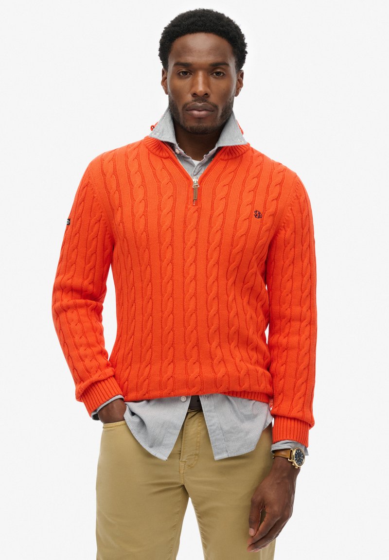Pomarańczowy sweter z dzianiny warkoczyków z pół-zamkiem, noszony na jasnoniebieskiej koszuli z guzikami. Posiada ściągacze na rękawach i u dołu. Prosty detal z logo.