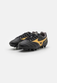 Zapatillas de fútbol negras hechas de cuero sintético con acentos amarillos, que cuentan con una parte superior texturizada, diseño de cordones y suela de goma moldeada con tacos.