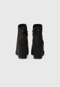 Juodos suede kulkšnies aulinukai su masyviu kulnu, turintys užtrauktuką ir apvalią metalinę detalę gale. Lygi tekstūra.